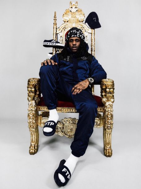 The rapper sitting on a golden throne looking chair in blue jacket and sweatpants with flipflops with 'FTP' written on it. alongside shoes and a hat hanging from the chair. 
